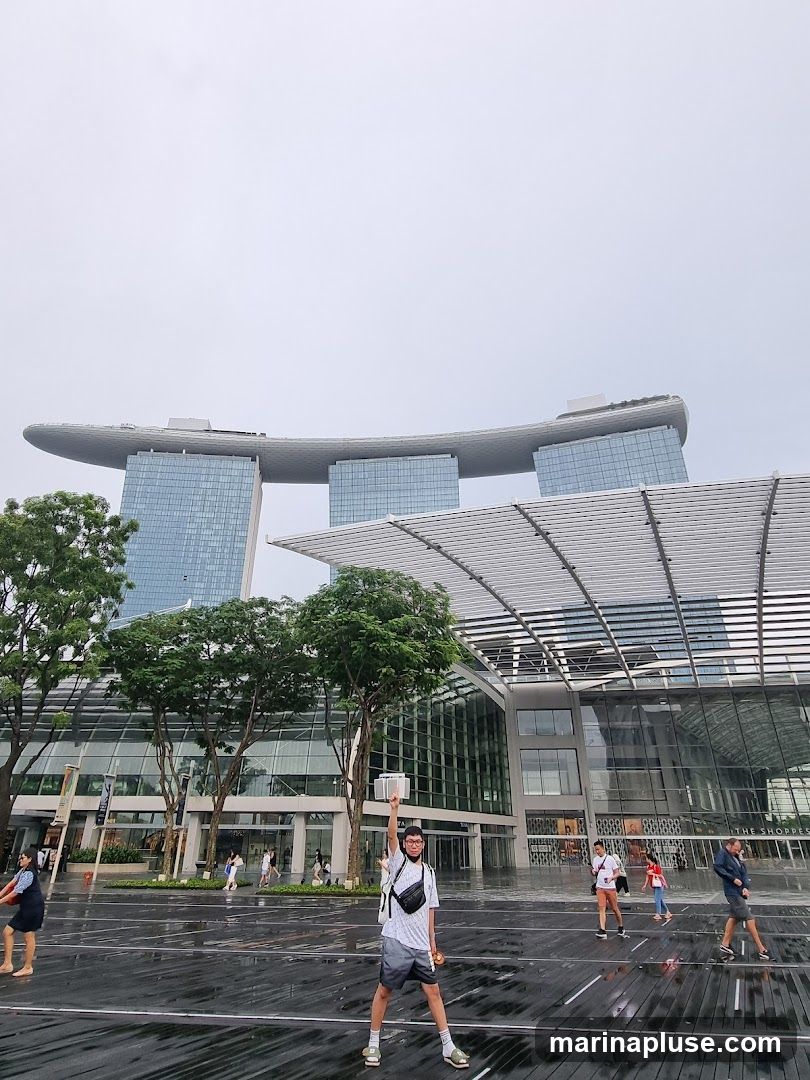 A person stands in the foreground with their arm raised, holding a white object. In the background, the iconic Marina Bay Sands hotel and its connected shopping complex are visible. Several other people are walking around the wet plaza in front of the buildings. The sky is overcast.