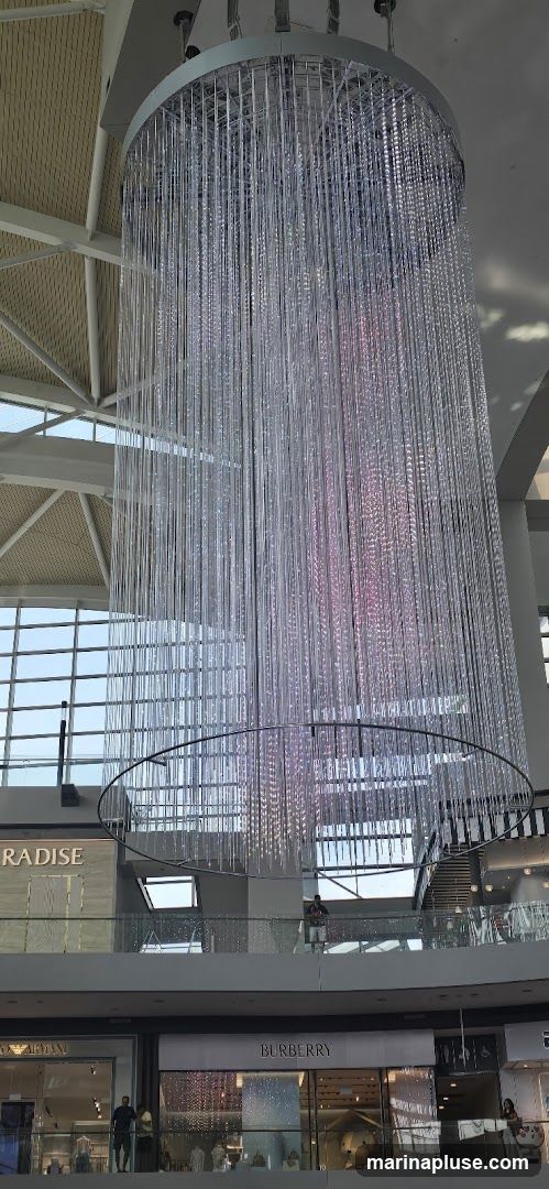 A large, cylindrical chandelier made of clear strands hangs from the ceiling of a mall. The strands are illuminated with pink and blue lights. Below the chandelier, storefronts are visible on two levels. Signs for "PARADISE", "ARMANI", and "BURBERRY" can be seen.
