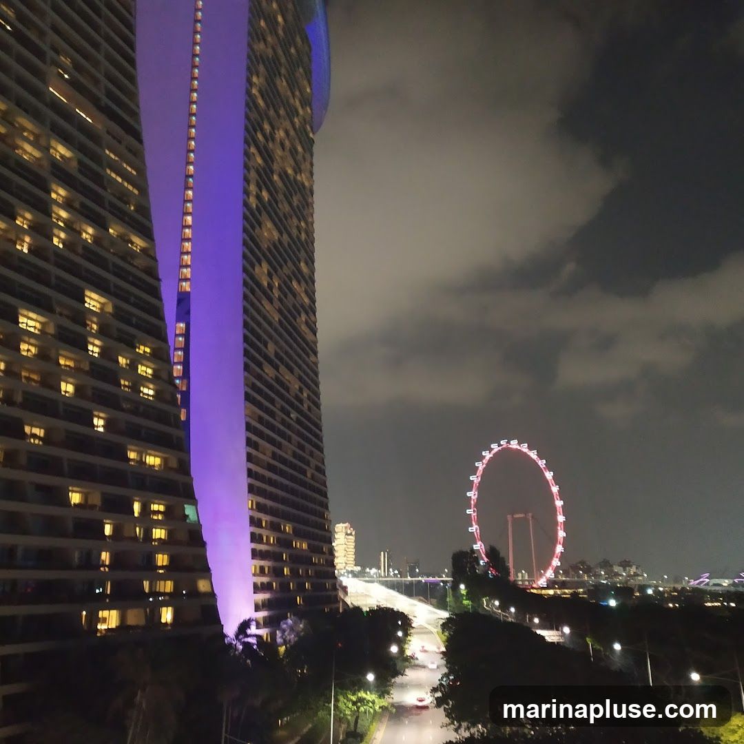 A nighttime view of a city skyline featuring two tall buildings, one illuminated with purple light. In the distance, a Ferris wheel glows red against a cloudy sky. A road with cars is visible in the foreground, lined with trees and streetlights.