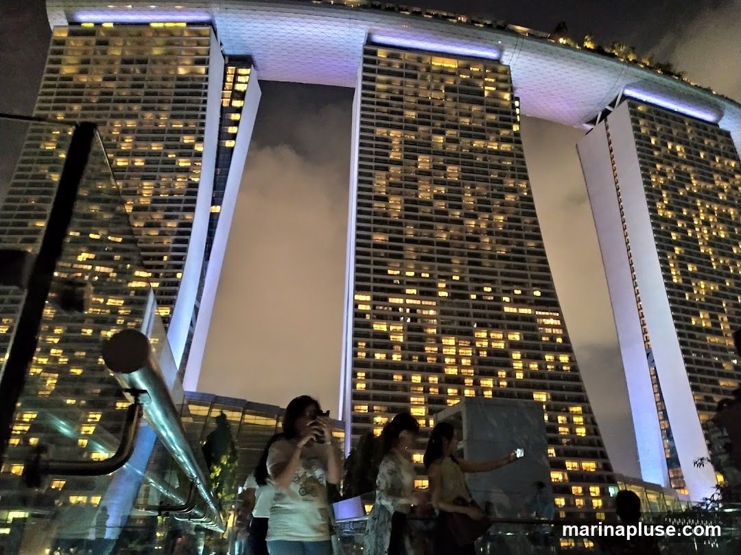A nighttime view of the iconic Marina Bay Sands hotel in Singapore, with its three towers connected by a rooftop deck. The building is illuminated with many lights, and the sky is dark with some clouds. Several people are visible in the foreground, appearing to take photos or selfies.