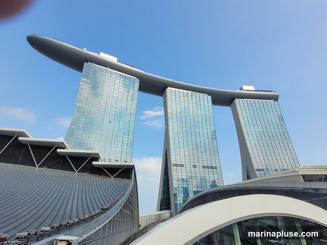 A low-angle shot of the Marina Bay Sands hotel in Singapore, with its distinctive three towers topped by a boat-shaped structure. The sky is clear blue with a few wispy clouds. The architecture is modern and sleek, with glass facades reflecting the sky.