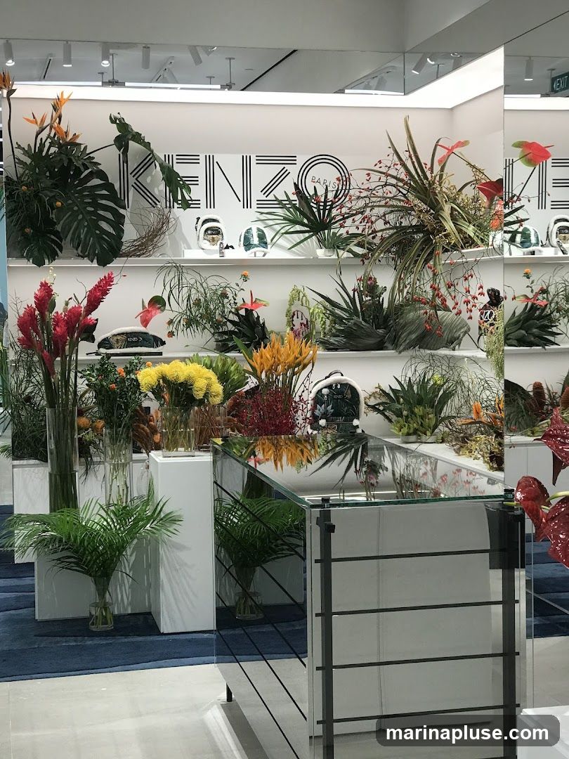 A display of various flowers and plants arranged on shelves and in vases. The KENZO PARIS logo is visible on the back wall. A mirrored counter is in the foreground.