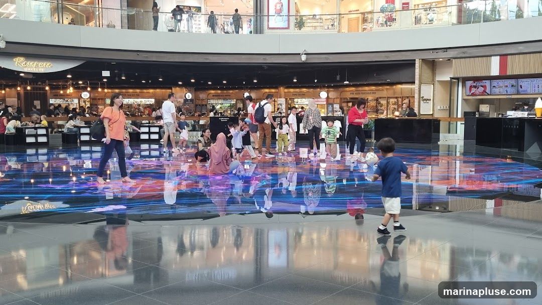 A group of people, including adults and children, are walking and standing in a large, open indoor space. The floor has a vibrant, digital display that reflects the surroundings. In the background, there are various shops and food establishments. One shop has a sign that reads 'Rasapura Masters'. Another area shows a menu board with items like 'RAMEN', 'JAPANESE SOBA', and 'ICE CREAM'.