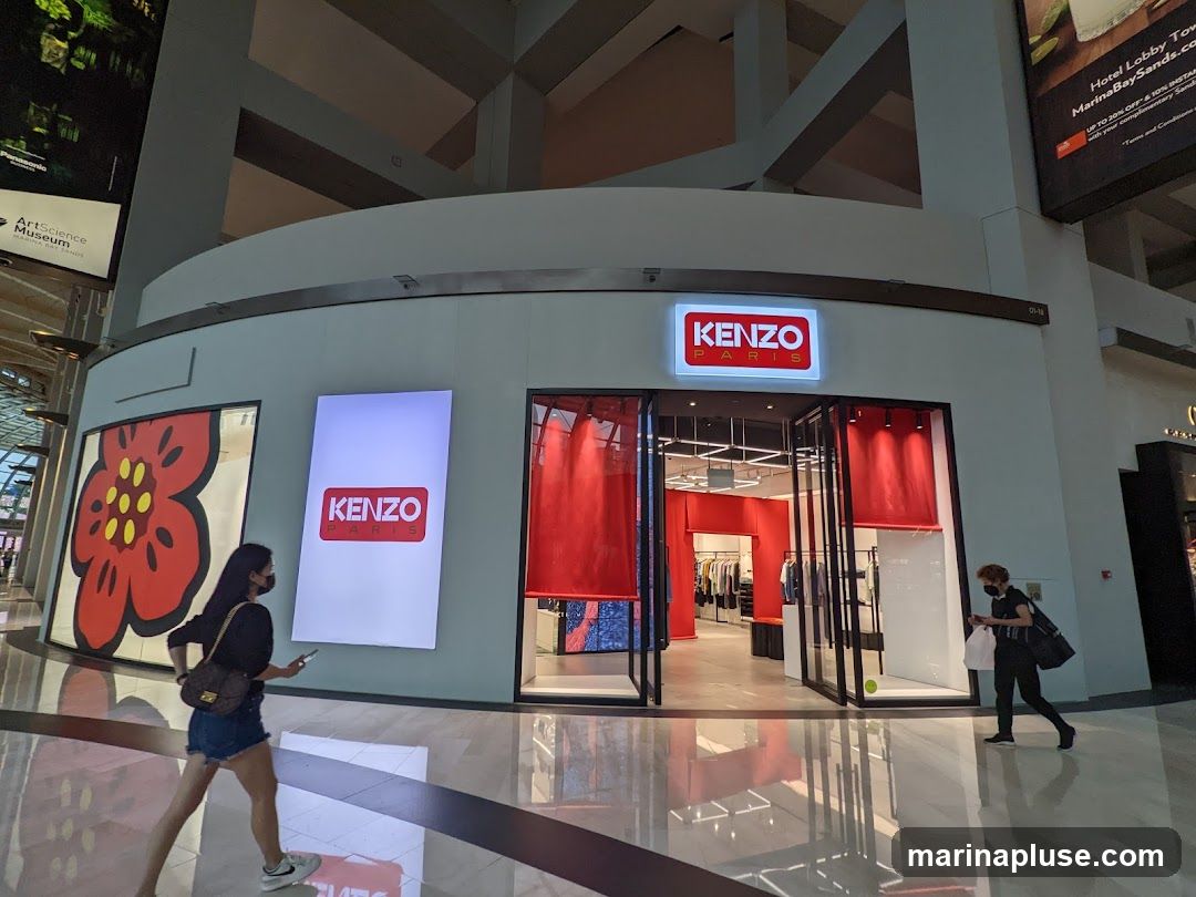 A well-lit storefront for Kenzo Paris, featuring the brand's logo prominently above the entrance. The interior of the store is visible through large glass doors, displaying clothing racks and red drapery. To the left of the entrance, a large digital display shows a stylized red flower, and another vertical display shows the Kenzo Paris logo. Two individuals are walking past the storefront.