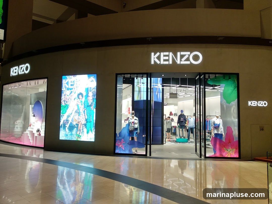 The exterior of a Kenzo store with large glass windows and doors. The Kenzo logo is prominently displayed in white illuminated lettering above the entrance and on the side of the building. Inside, mannequins are dressed in fashionable clothing, and displays showcase various products. Digital screens on the facade feature vibrant imagery and advertisements.