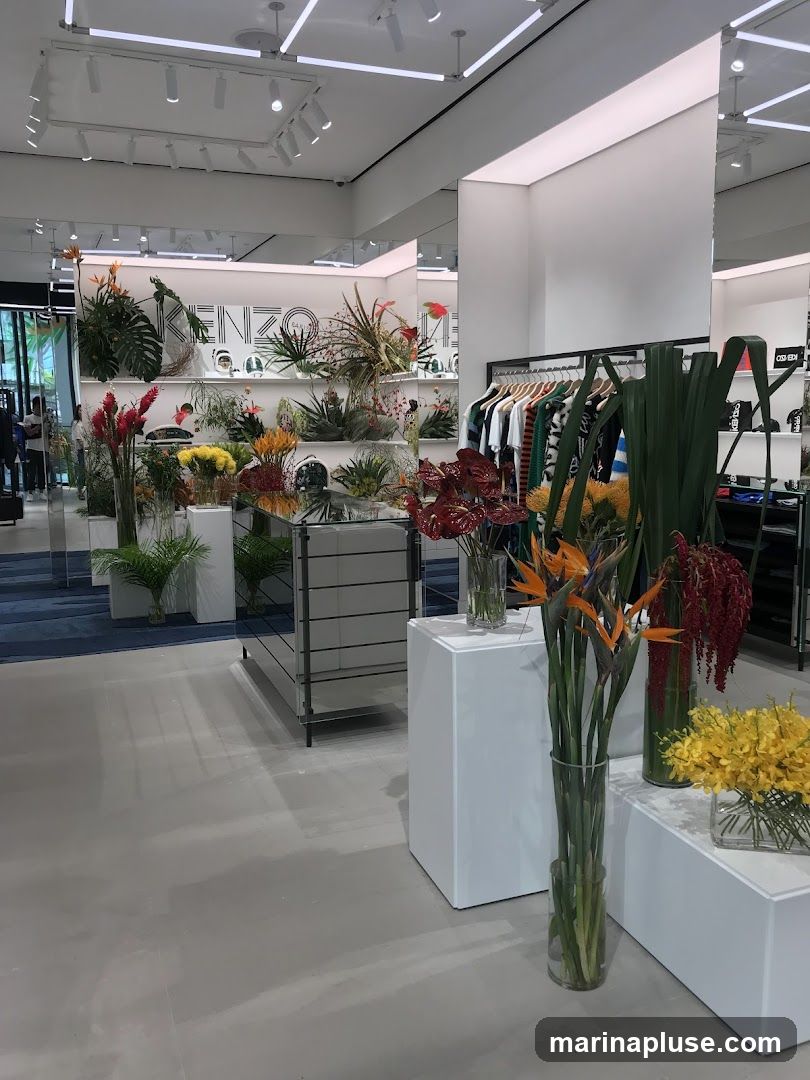 The image shows the interior of a store, likely a boutique or department store, with a focus on floral arrangements and clothing displays. In the background, a wall features the KENZO logo prominently, surrounded by various plants and decorative items. Racks of clothing, including t-shirts and sweaters, are visible on the right side of the image. Several large vases filled with vibrant flowers, such as bird of paradise and red anthuriums, are placed throughout the store, adding a touch of color and life to the modern, minimalist setting. The floor is a light gray, and the ceiling is equipped with track lighting. Mirrors are used to enhance the sense of space.