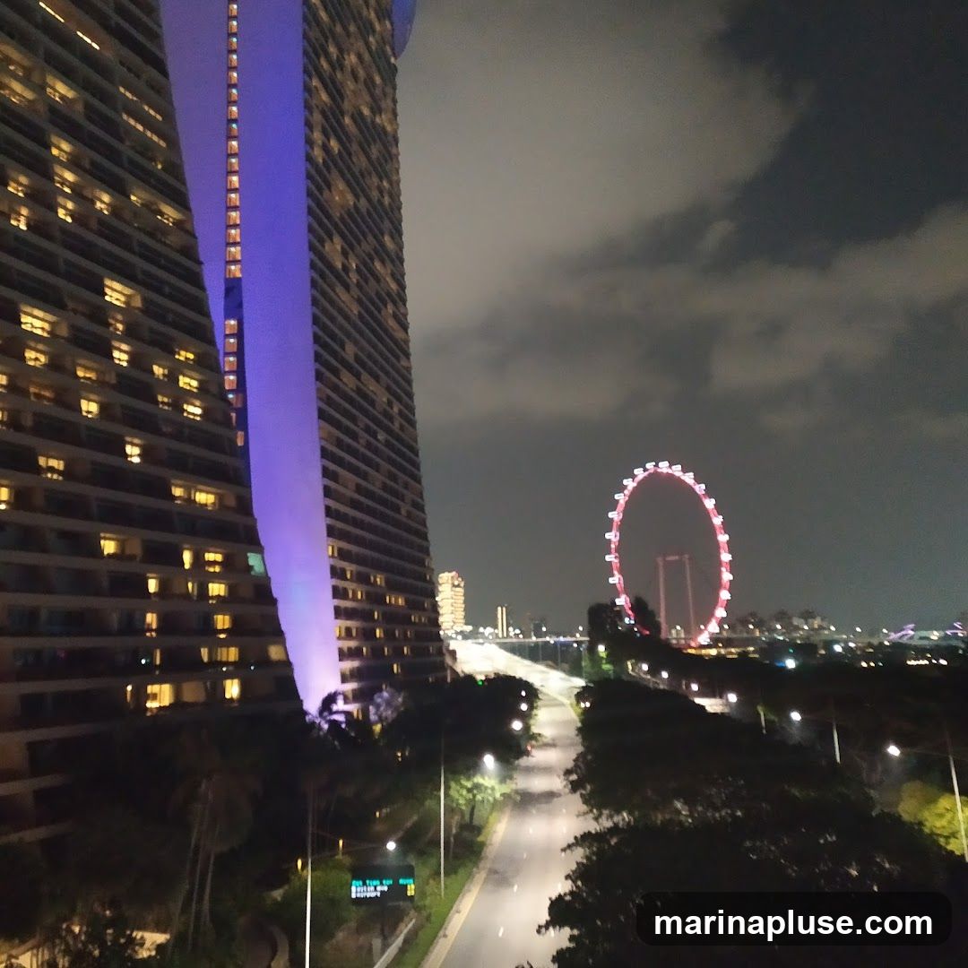A nighttime view of the Marina Bay Sands hotel with its distinctive architecture illuminated by purple light. To the right, the Singapore Flyer Ferris wheel is lit up in red. A road runs through the foreground, flanked by trees and streetlights. The sky is cloudy.