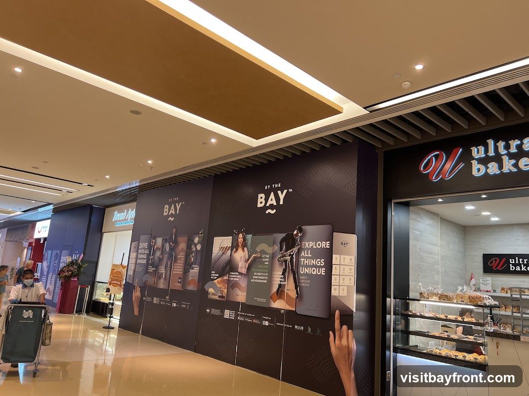 A view of a bakery with its display cases filled with pastries and bread. Above the entrance, the bakery's name is visible. To the left of the bakery, a large advertisement panel displays various images and text related to "BY THE BAY," promoting exploration of unique things. A person in a mask is seen pushing a shopping cart in the foreground.