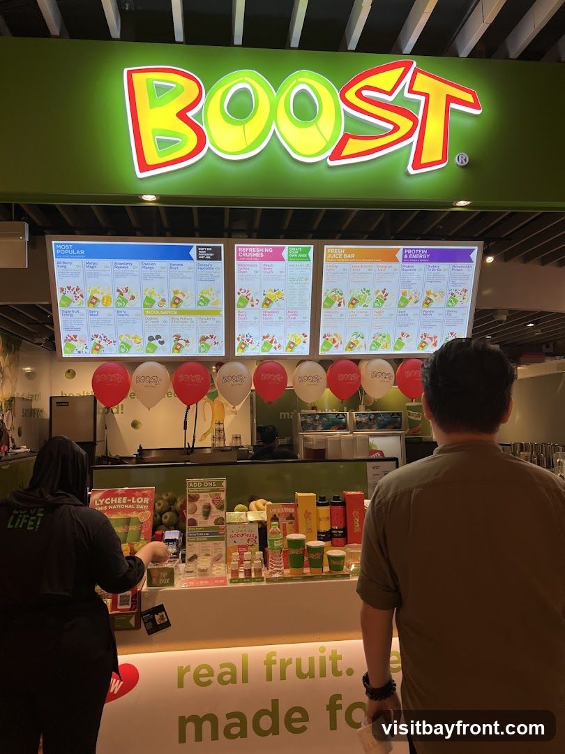 A Boost Juice Bar with a large illuminated sign and menu boards displaying various drinks. Two customers are visible, one ordering at the counter and another standing in front of it. Red and white balloons are strung across the top of the counter.