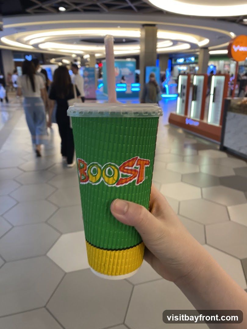 A hand holds a green and yellow corrugated paper cup with the "BOOST" logo visible. The cup has a white lid and a pink straw. The background shows a blurred indoor shopping mall with people walking and illuminated displays.
