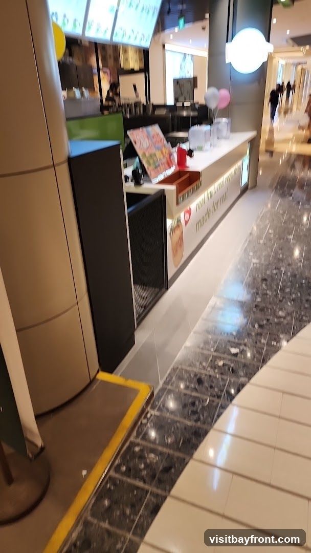 A juice bar counter with a menu board above and a display of fresh fruits. The counter has a white surface with a wooden trim. There are several clear containers on the counter, possibly for ingredients or drinks. A red dispenser is also visible. A sign on the side of the counter reads "real fruit, real vegetables, made for real." The floor is tiled with a dark, speckled pattern. In the background, people are walking through a mall.