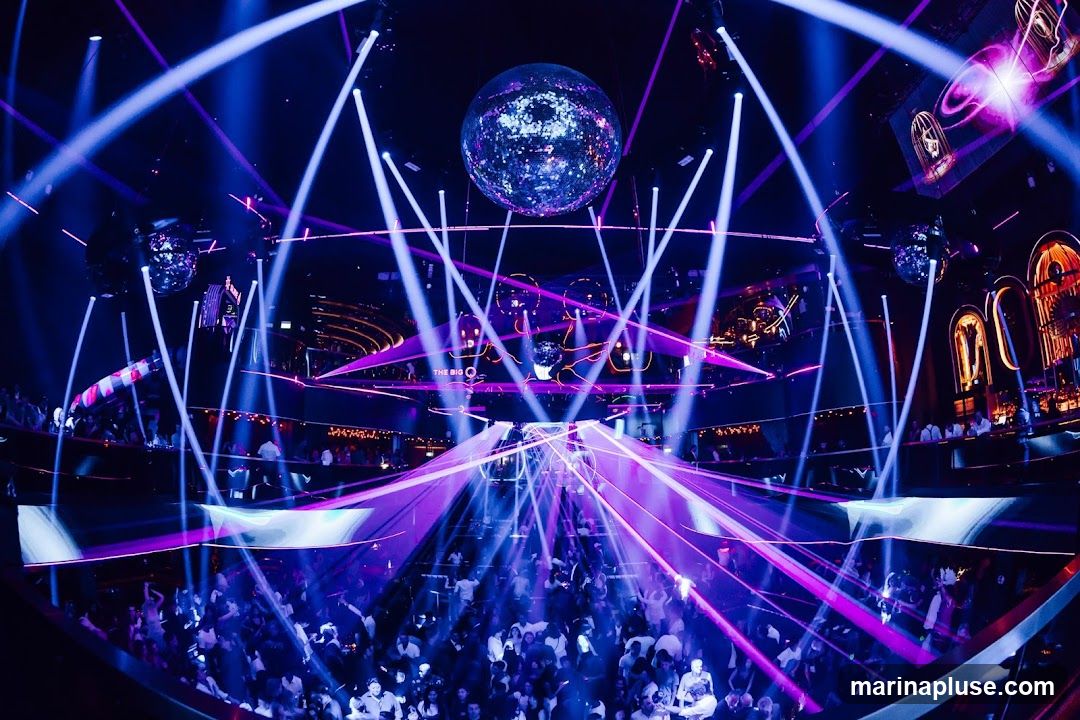 A crowded nightclub is illuminated by a large disco ball and numerous blue and purple laser beams. People are dancing on multiple levels of the venue.
