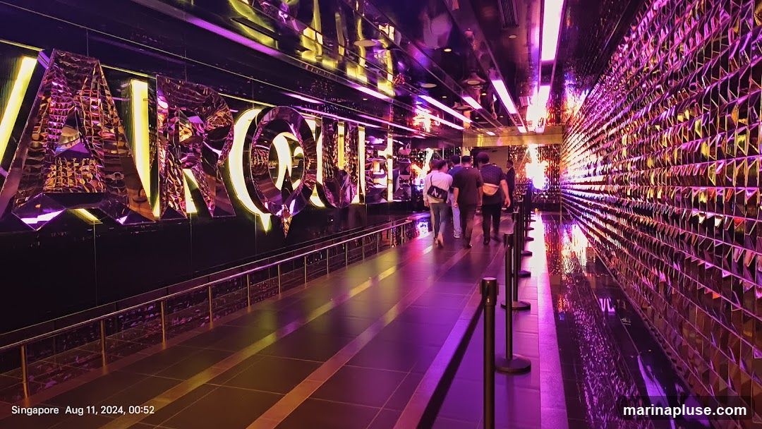 A walkway with large, illuminated, mirrored letters spelling out "MARQUEE" on the left wall. The walkway is lined with stanchions and leads towards a group of people. The right wall is covered in a mosaic of reflective tiles, creating a dazzling effect with purple and pink lighting. The ceiling is also mirrored, reflecting the lights and the overall ambiance of the space.