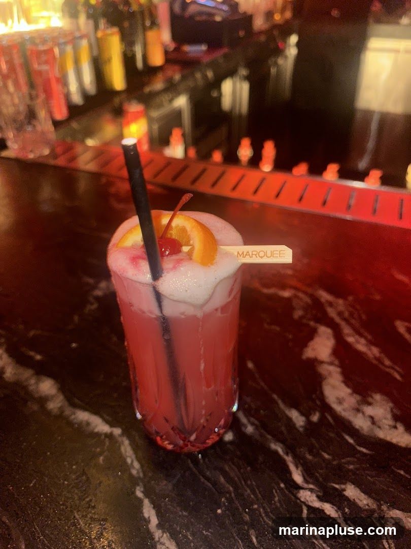 A pink cocktail with foam, an orange slice, and a cherry sits on a dark marble bar. A black straw is in the drink, and a small wooden pick with the word "MARQUEE" is inserted into the garnish. In the background, bottles and cans are visible on shelves behind the bar, with red lighting illuminating the area.