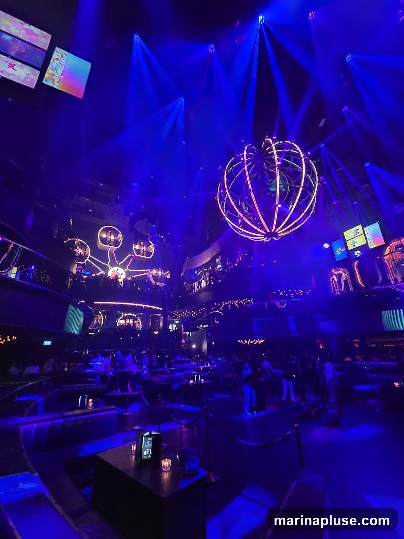A wide shot of a nightclub interior illuminated by vibrant blue lights. A large, spherical light fixture with pink neon accents hangs from the ceiling, surrounded by numerous spotlights casting beams across the room. Multiple levels of seating areas with tables and banquettes are visible, occupied by patrons. Screens displaying colorful graphics are mounted on the walls.