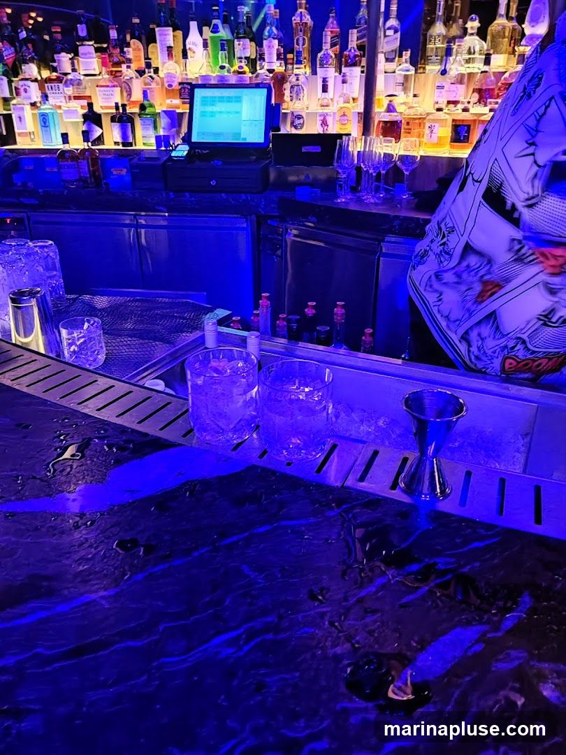 A bar counter with bottles of alcohol displayed on shelves behind it. There are glasses with ice and drinks on the counter, along with a jigger and a POS system. The lighting is predominantly blue.