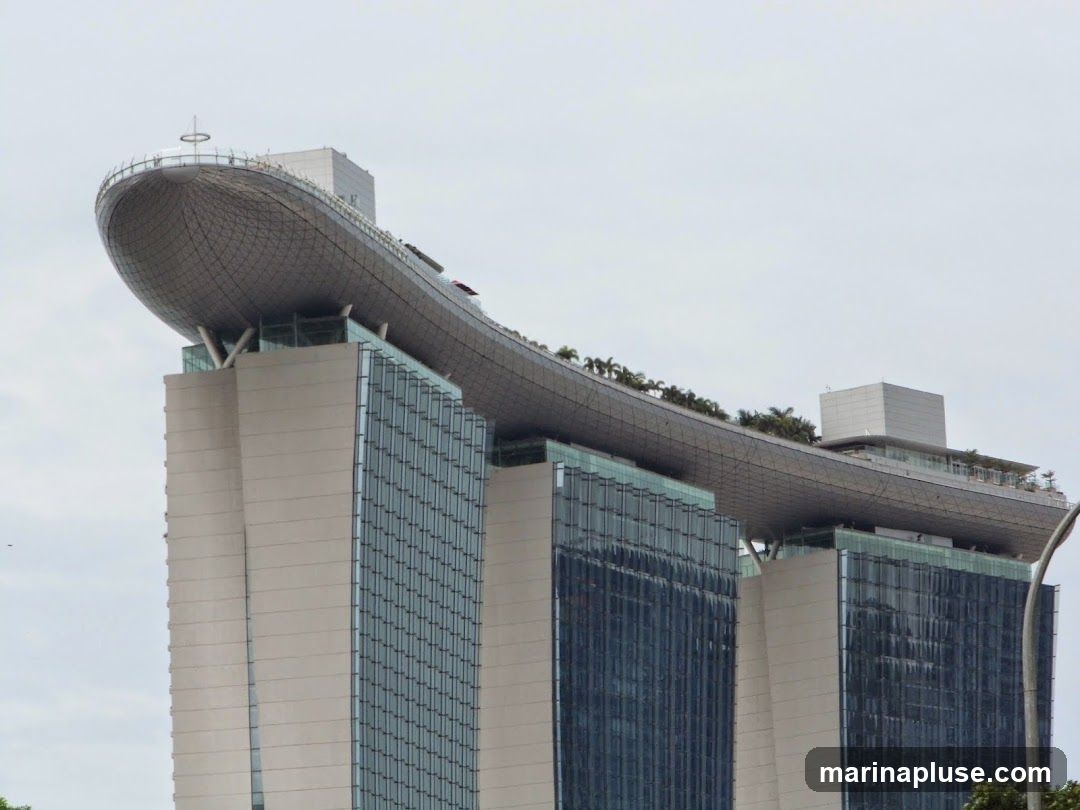 A view of the Marina Bay Sands hotel in Singapore, focusing on the iconic Skypark observation deck that spans across the three towers. The Skypark features a distinctive boat-like shape and is topped with a circular railing and a small antenna. Lush greenery is visible along the edge of the Skypark, suggesting a garden or landscaped area. The hotel towers themselves are characterized by their modern architecture, with glass facades reflecting the sky and beige concrete sections. The sky is overcast, providing a soft, diffused light.