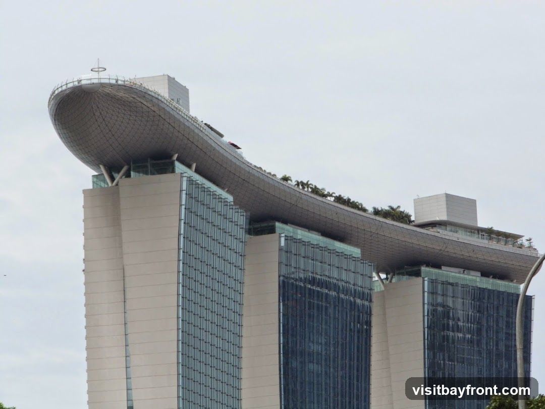 A view of the Marina Bay Sands hotel in Singapore, focusing on the iconic Skypark observation deck that spans across the three towers. The Skypark features a distinctive boat-like shape and is topped with a circular railing and a small antenna. Lush greenery is visible along the edge of the Skypark, suggesting a garden or landscaped area. The hotel towers themselves are characterized by their modern architecture, with glass facades reflecting the sky and beige concrete sections. The sky is overcast, providing a soft, diffused light.