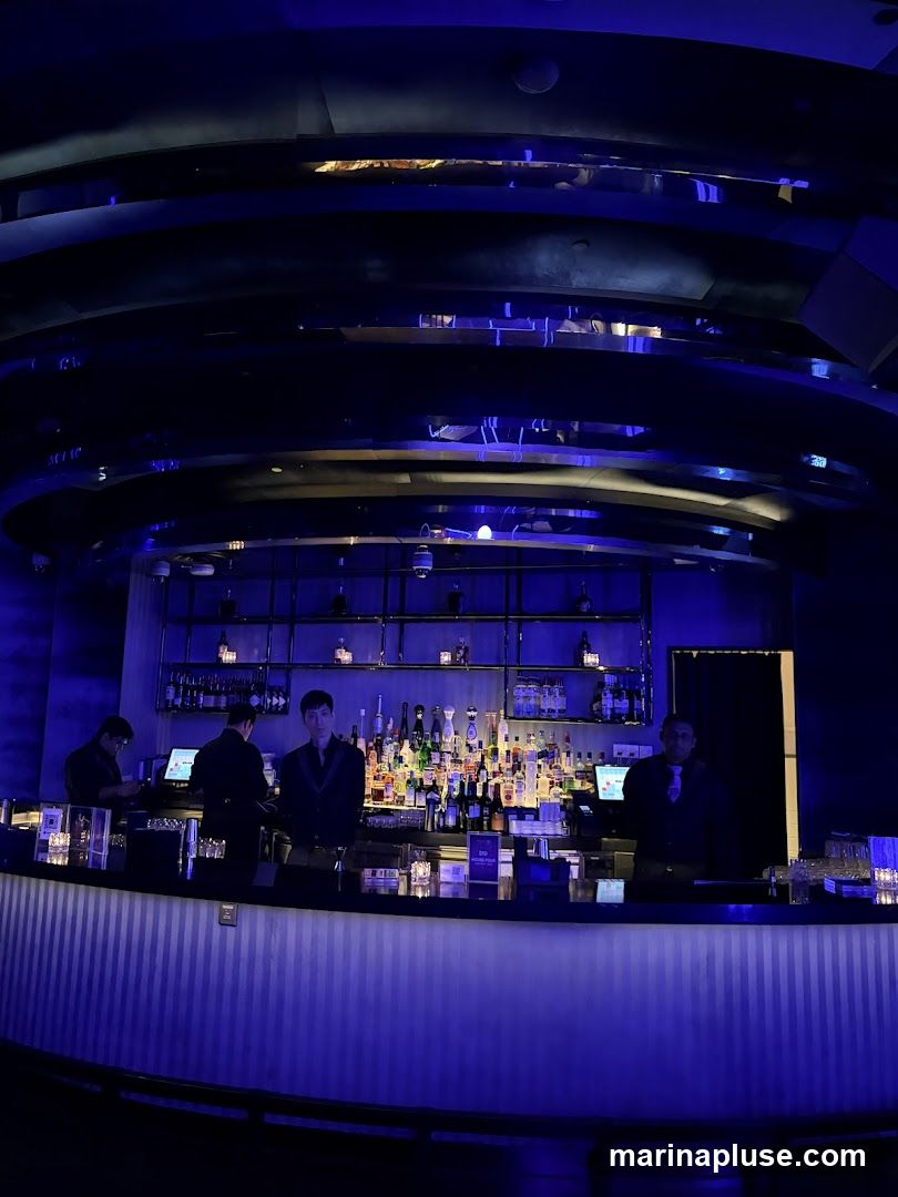 A dimly lit bar interior with a long, curved bar illuminated by blue lights. Bartenders are visible behind the bar, which is stocked with numerous bottles of liquor. The ceiling features a modern, multi-layered design with reflective surfaces.
