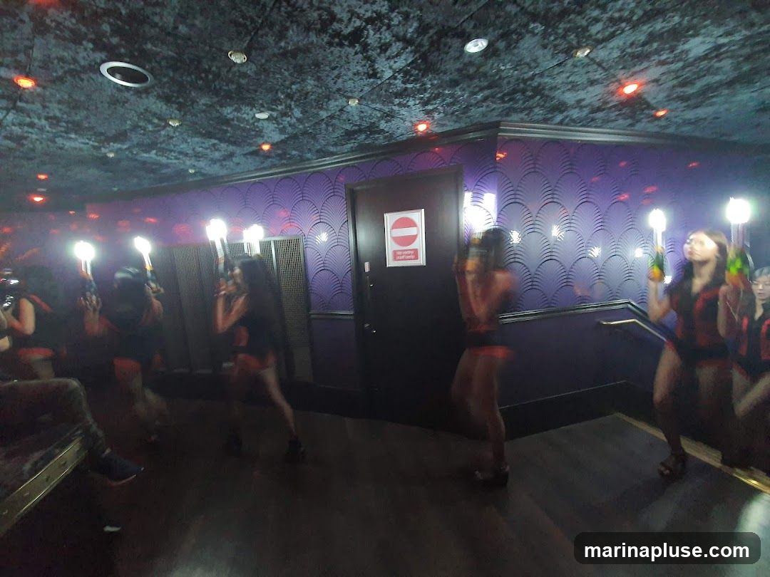 A group of women in matching outfits are holding illuminated devices and bottles, possibly champagne, in a dimly lit nightclub. The walls are decorated with a purple, fan-like pattern, and the ceiling has textured dark material with recessed lighting. A door with a "no entry" sign is visible on the left.