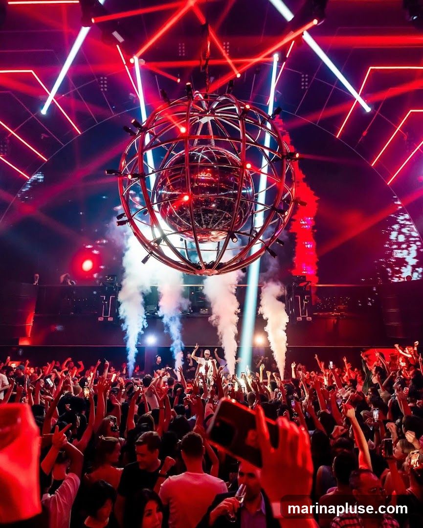 A crowded nightclub with a DJ on stage, a large spherical light fixture hanging from the ceiling, and red and white laser lights illuminating the room. Smoke machines are active, and the audience is dancing with their hands in the air.