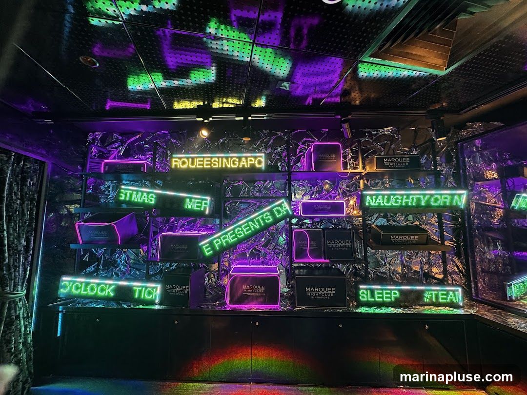 A display wall with shelves featuring various items and neon signs. The signs read "ROUEESINGAPO", "STMAS MER", "E PRESENTS DA", "NAUGHTY OR N", "S'CLOCK TICH", and "SLEEP #TEA". Several boxes with the "MARQUEE NIGHTCLUB SINGAPORE" logo are also visible on the shelves. The background has a metallic, crinkled texture, and the ceiling is illuminated with green and purple lights.