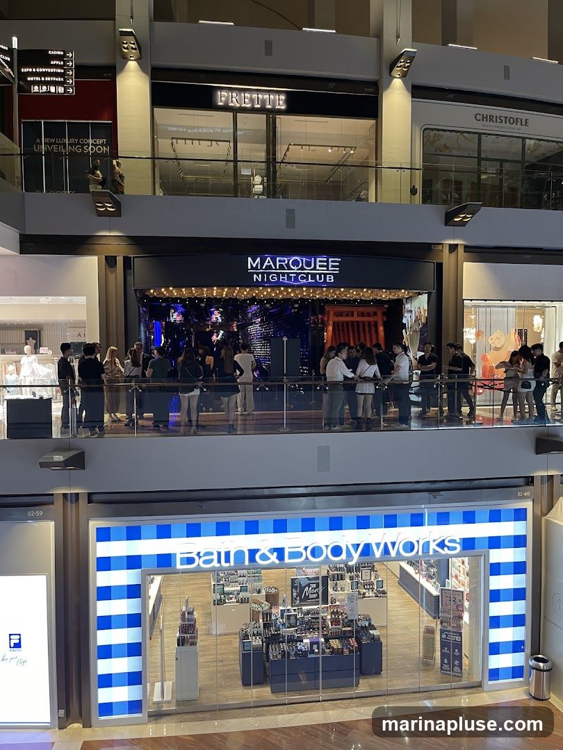 An interior view of a multi-level shopping mall. On the top level, signs indicate directions to CASINO, EXPO & CONVENTION, HOTEL & SKYPARK, and a new luxury concept unveiling soon. Further along, the storefronts for FRETTE and CHRISTOFLE are visible. On the middle level, the entrance to MARQUEE NIGHTCLUB is prominent with a crowd gathered. On the lower level, the brightly lit storefront of Bath & Body Works is visible, displaying various products inside.