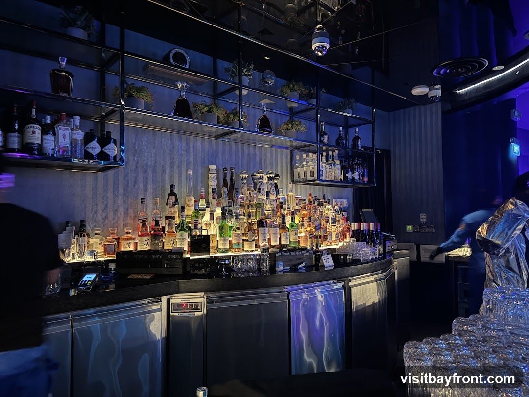 A well-stocked bar with numerous bottles of alcohol displayed on shelves and the counter. The lighting is dim and blue, creating a sophisticated atmosphere. There are stacks of clean glasses in the foreground, ready for use. A person in a shiny silver jacket is visible in the background, appearing to be working behind the bar.