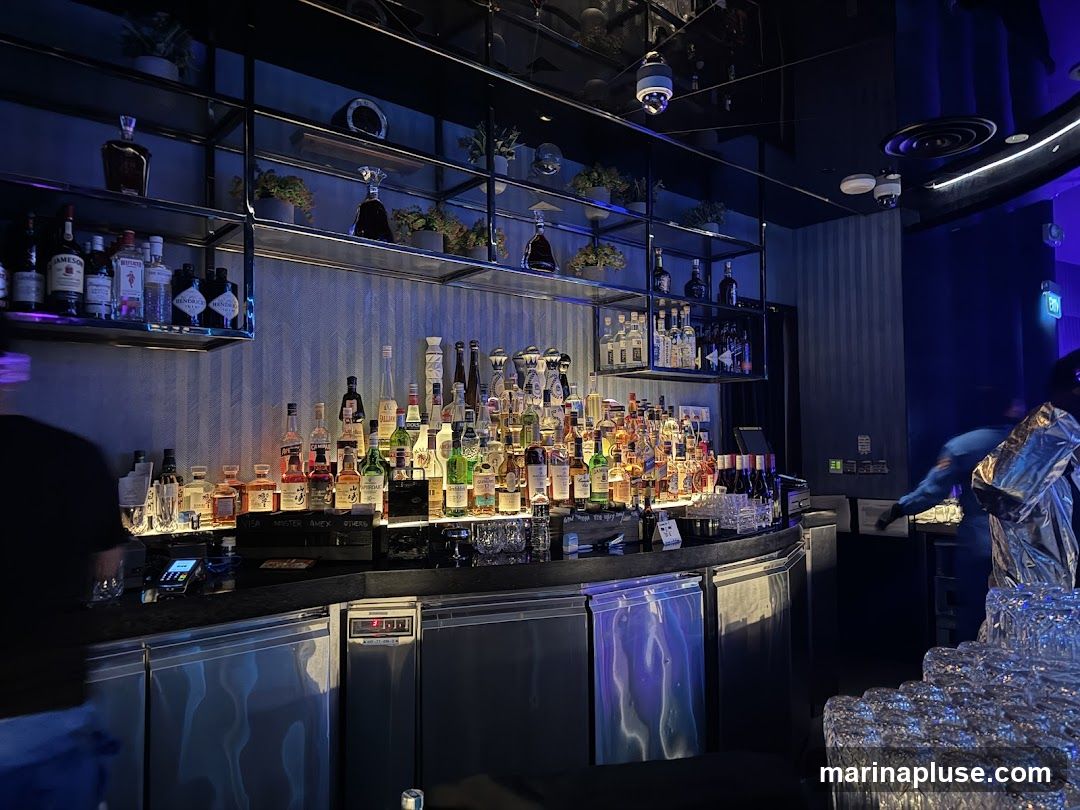 A well-stocked bar with numerous bottles of alcohol displayed on shelves and the counter. The lighting is dim and blue, creating a sophisticated atmosphere. There are stacks of clean glasses in the foreground, ready for use. A person in a shiny silver jacket is visible in the background, appearing to be working behind the bar.