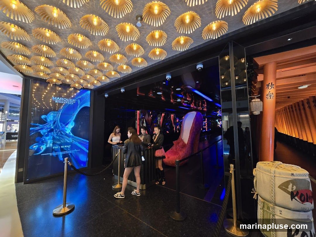 The entrance to Marquee Singapore features a large digital screen displaying abstract blue imagery and the text "MARQUEE SINGAPORE FRI 15 MAY". Several people are standing near the entrance, and a velvet rope guides visitors. The ceiling is adorned with numerous decorative light fixtures. To the right, there is an orange-themed area with Japanese calligraphy and stacked sake barrels. A large, plush red high-heeled shoe is visible inside the venue.