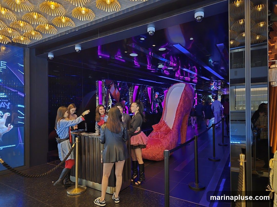 A group of people are gathered at a reception desk inside a dimly lit venue. The walls are dark and reflective, with purple lighting accents. A large, plush red high-heeled shoe is prominently displayed as a decorative element. A digital screen on the left shows graphics and text, possibly advertising an event. The overall ambiance suggests a nightclub or entertainment venue.