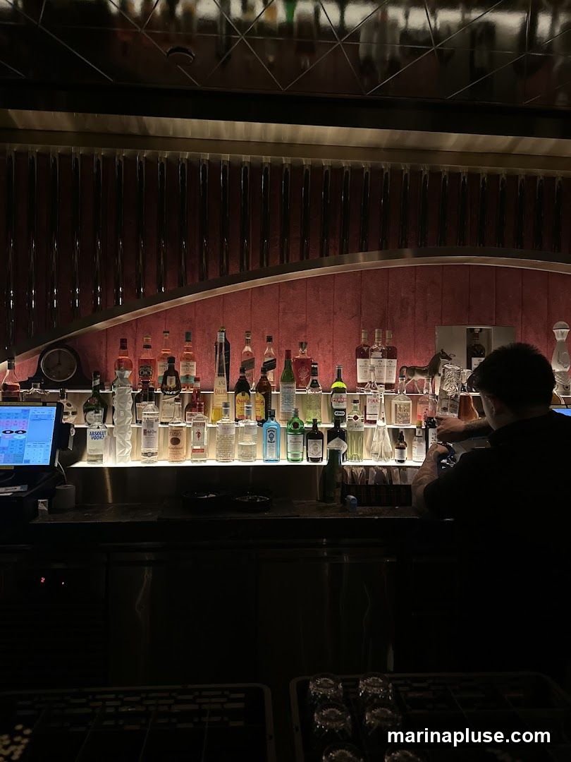 A dimly lit bar interior with a bartender preparing drinks behind a counter stocked with numerous bottles of alcohol. The bottles are arranged on shelves illuminated from below, creating a dramatic effect. The background features a textured, dark red wall with vertical ribbed detailing and a reflective ceiling. A POS system is visible on the left side of the counter.