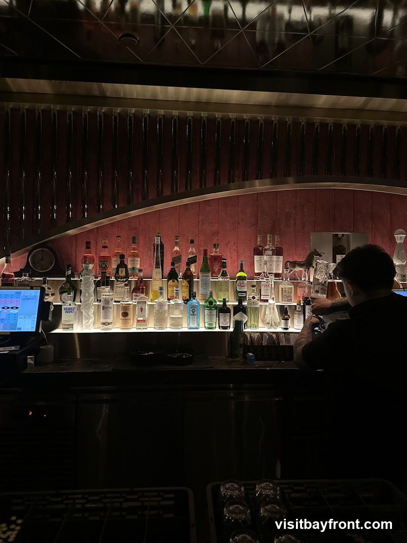 A dimly lit bar interior with a bartender preparing drinks behind a counter stocked with numerous bottles of alcohol. The bottles are arranged on shelves illuminated from below, creating a dramatic effect. The background features a textured, dark red wall with vertical ribbed detailing and a reflective ceiling. A POS system is visible on the left side of the counter.