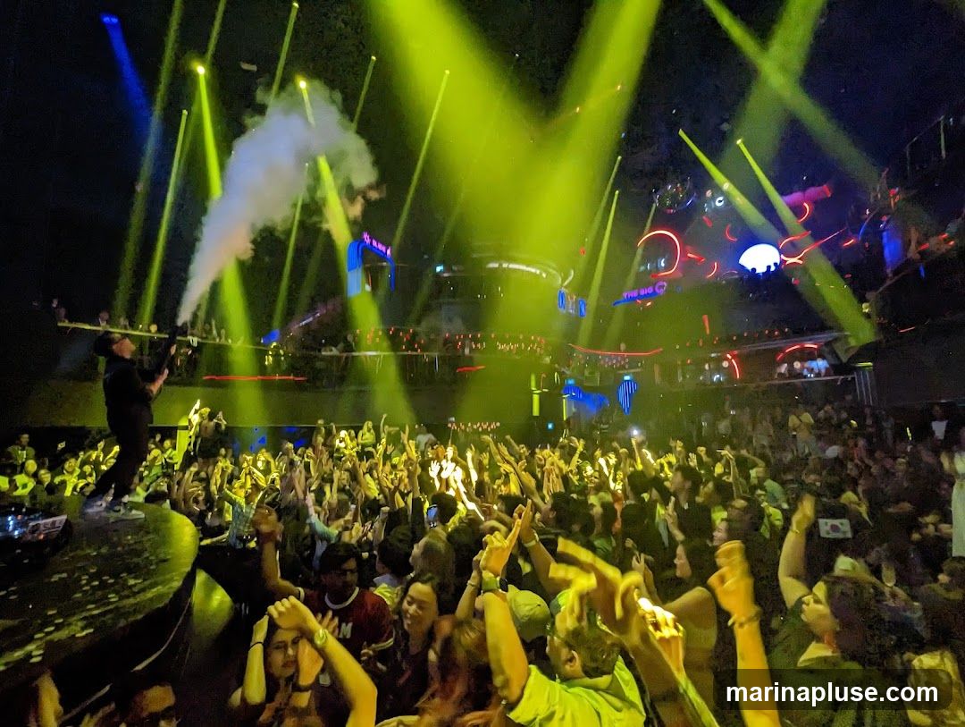 A DJ on a platform is spraying smoke into a crowded nightclub. The crowd is enthusiastically dancing and raising their hands. Bright yellow and blue lights illuminate the scene, with neon rings and a disco ball visible in the background. The sign 'THE BIG O' is visible.