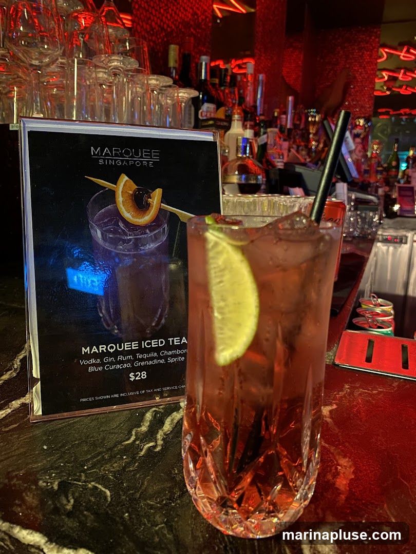 A close-up of a Marquee Iced Tea cocktail with a lime wedge and a straw, next to a menu displaying the drink's ingredients and price. The background shows a bar with bottles and glassware.