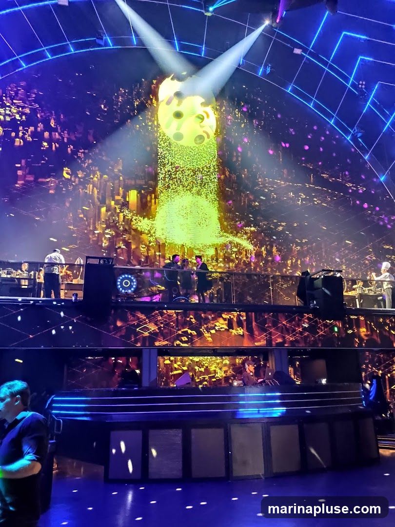 A vibrant concert stage is illuminated with dramatic lighting. A large, yellow, circular structure with cutouts hangs from the ceiling, emitting a bright green light that cascades down like a waterfall of glitter. The background features a digital cityscape projection in shades of gold and orange. Several performers, including DJs and musicians, are visible on different levels of the stage. The foreground shows a dimly lit area with a few audience members.