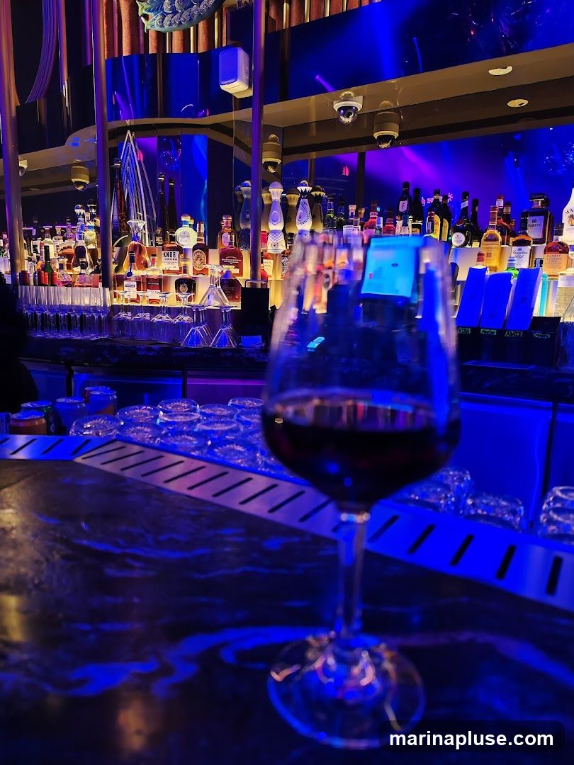 A wine glass filled with red wine sits in the foreground on a dark, reflective bar top. Behind it, a well-stocked bar is illuminated with vibrant blue and purple lights. Numerous bottles of liquor are displayed on shelves, with some bottles featuring distinctive labels. In the background, decorative elements and lighting contribute to a sophisticated ambiance.