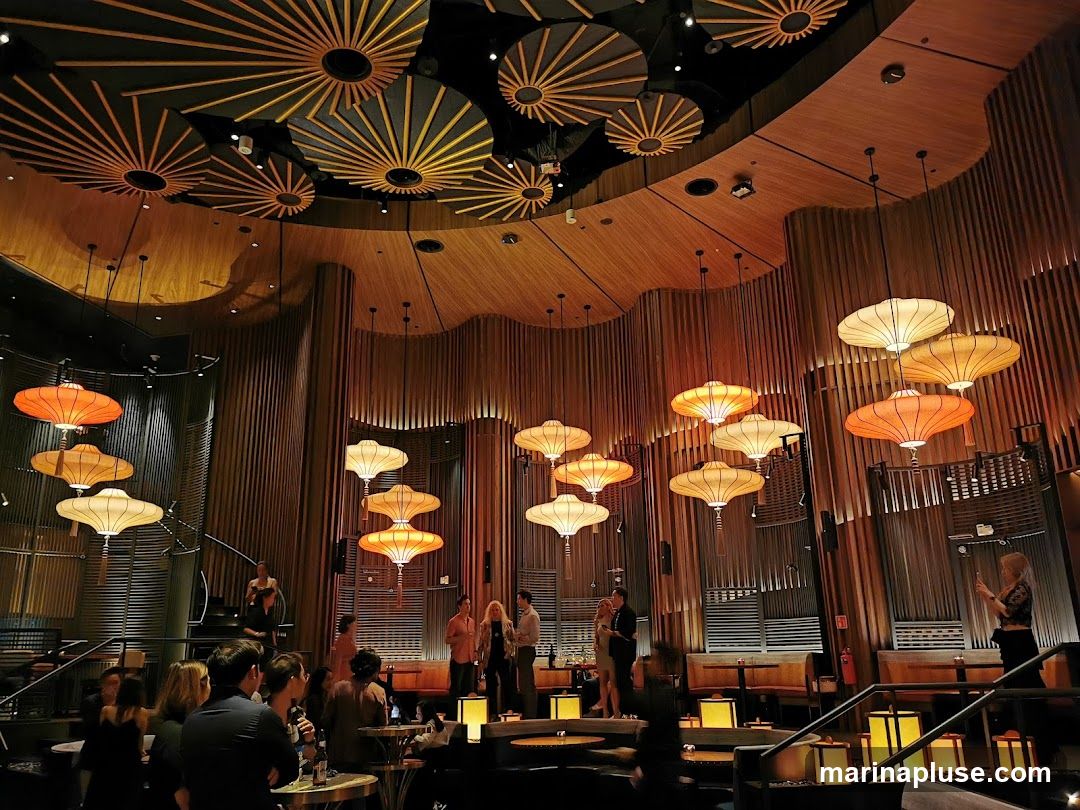 The interior of a restaurant with a distinctive ceiling design featuring radial patterns. Numerous large, tiered lanterns in orange and cream hues hang from the ceiling, illuminating the space. The walls are paneled with vertical wooden slats, creating a warm and textured ambiance. Several groups of people are seated at tables, some engaged in conversation, while others appear to be dining. The overall atmosphere is sophisticated and dimly lit, with a focus on the unique lighting fixtures and architectural details.