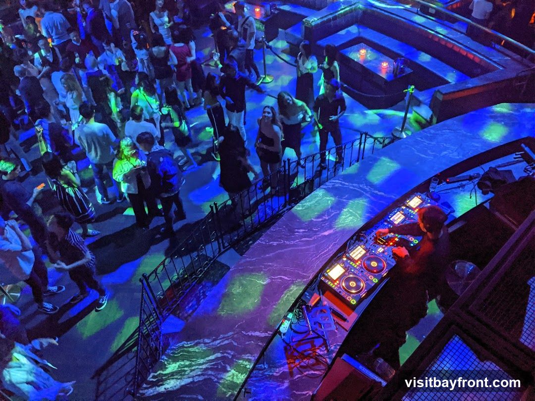 A crowded nightclub with a DJ booth in the foreground. The DJ is actively mixing music on a console with multiple decks and controls. The dance floor is filled with people, illuminated by vibrant blue and green lights that create a dynamic atmosphere. The lighting casts colorful patterns on the floor and walls, enhancing the energetic mood of the venue. In the background, there are elevated seating areas and architectural elements that contribute to the club's design.