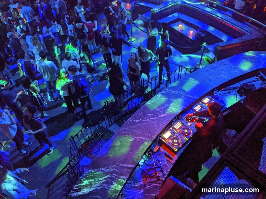A crowded nightclub with a DJ booth in the foreground. The DJ is actively mixing music on a console with multiple decks and controls. The dance floor is filled with people, illuminated by vibrant blue and green lights that create a dynamic atmosphere. The lighting casts colorful patterns on the floor and walls, enhancing the energetic mood of the venue. In the background, there are elevated seating areas and architectural elements that contribute to the club's design.