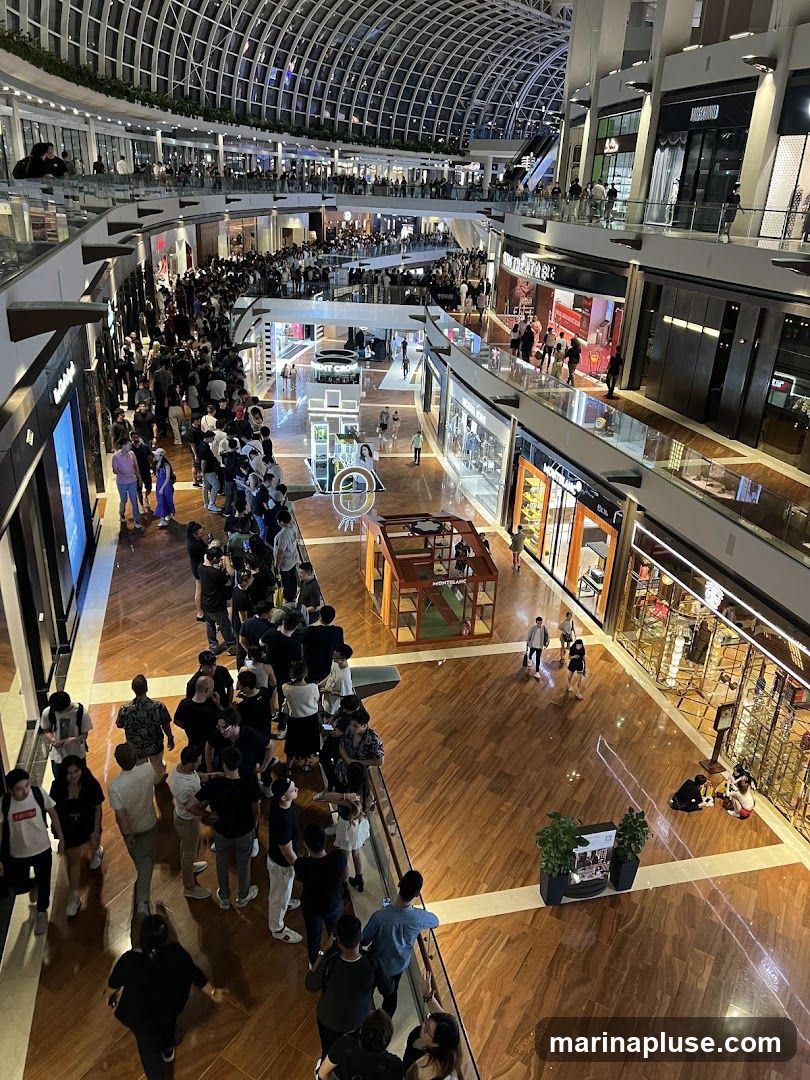A high-angle view of a busy, multi-level shopping mall interior with a large crowd of people queuing. The architecture features a distinctive domed glass ceiling and wooden flooring. Various storefronts are visible on different levels, with some displaying merchandise and signage. A display case with the Montblanc logo is present in the foreground.