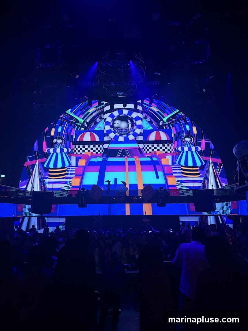 A vibrant and colorful stage setup with geometric patterns and lighting, featuring a DJ booth and a large disco ball. A crowd of people is visible in the foreground, silhouetted against the stage lights.
