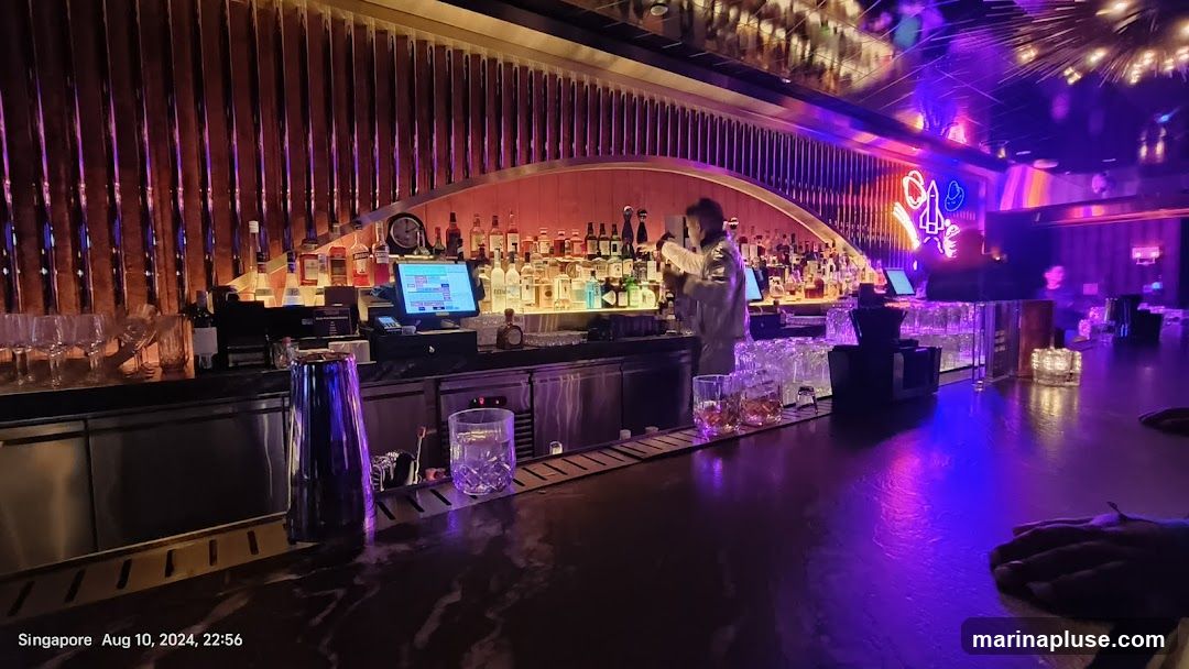 A dimly lit bar with a bartender preparing drinks. The bar is stocked with numerous bottles of alcohol, and glassware is neatly arranged. Neon lights, including a rocket and a cowboy hat, are visible in the background. The overall ambiance is modern and vibrant, with purple and pink lighting illuminating the space.