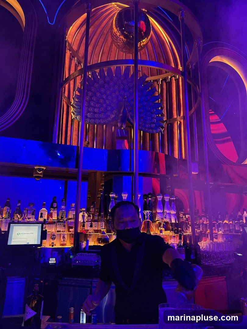 A bartender wearing a mask prepares a drink behind a bar stocked with numerous bottles of alcohol. Above the bar, a large decorative cage contains a peacock sculpture and a disco ball. The interior is lit with vibrant blue and purple lights, creating a nightclub atmosphere.