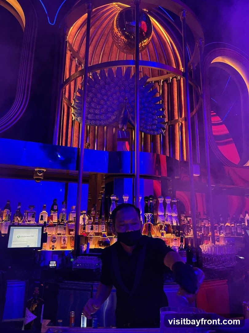 A bartender wearing a mask prepares a drink behind a bar stocked with numerous bottles of alcohol. Above the bar, a large decorative cage contains a peacock sculpture and a disco ball. The interior is lit with vibrant blue and purple lights, creating a nightclub atmosphere.