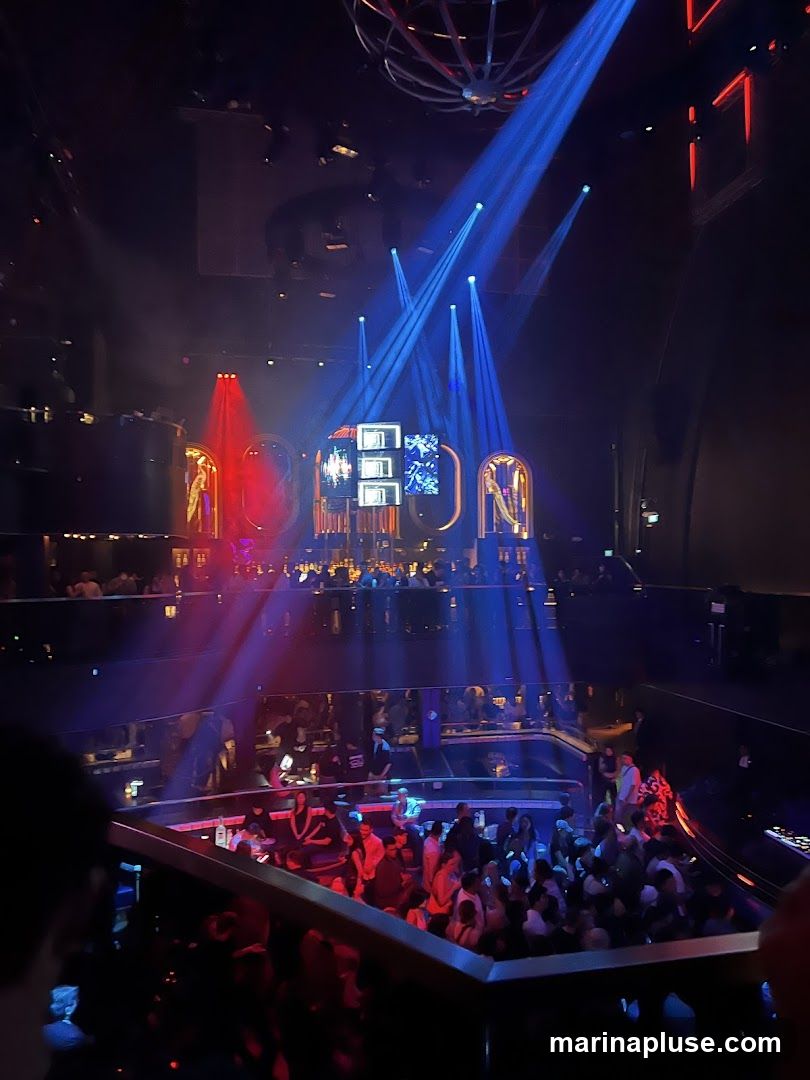 A vibrant nightclub scene with a stage featuring illuminated letters spelling out 'EON'. Blue and red spotlights cut through the smoky atmosphere, illuminating a large crowd gathered on multiple levels. People are seen socializing and enjoying the ambiance.