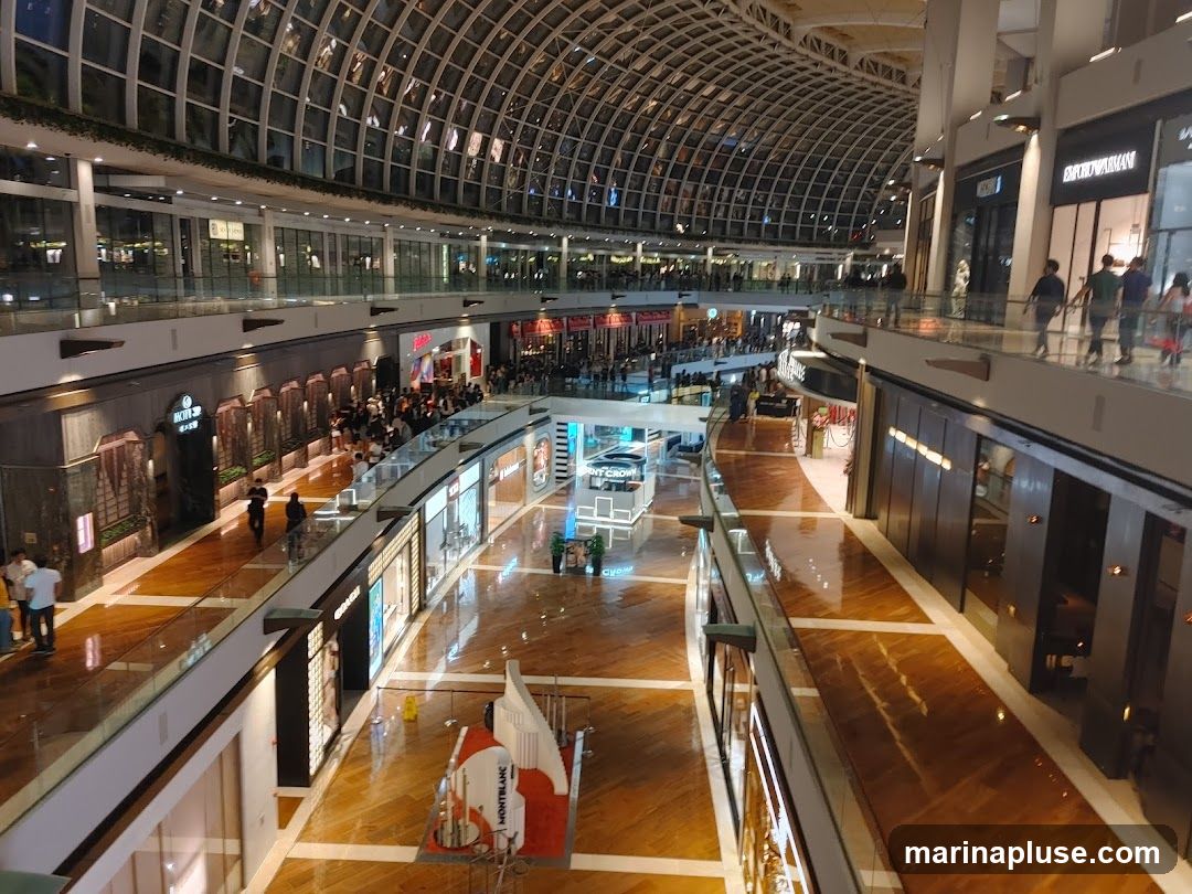 The image shows a wide, multi-level interior of a shopping mall with a distinctive arched glass ceiling. Numerous shops with illuminated storefronts line the walkways. People are seen browsing and walking along the polished wooden floors. In the foreground, a display featuring the Montblanc logo is visible.