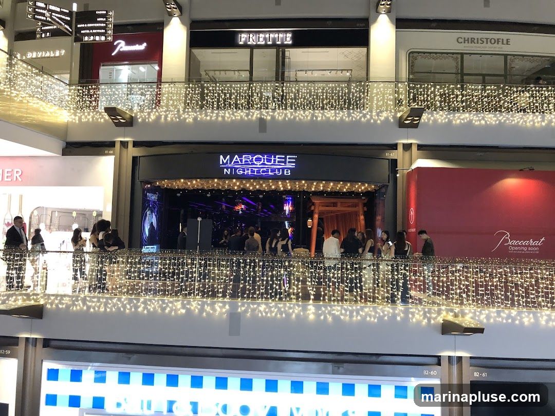 The exterior of a nightclub called Marquee Nightclub, with its name illuminated in neon blue above the entrance. People are gathered on a balcony in front of the club, which is decorated with strings of fairy lights. Other businesses, including Frette and Christofle, are visible on the upper levels.