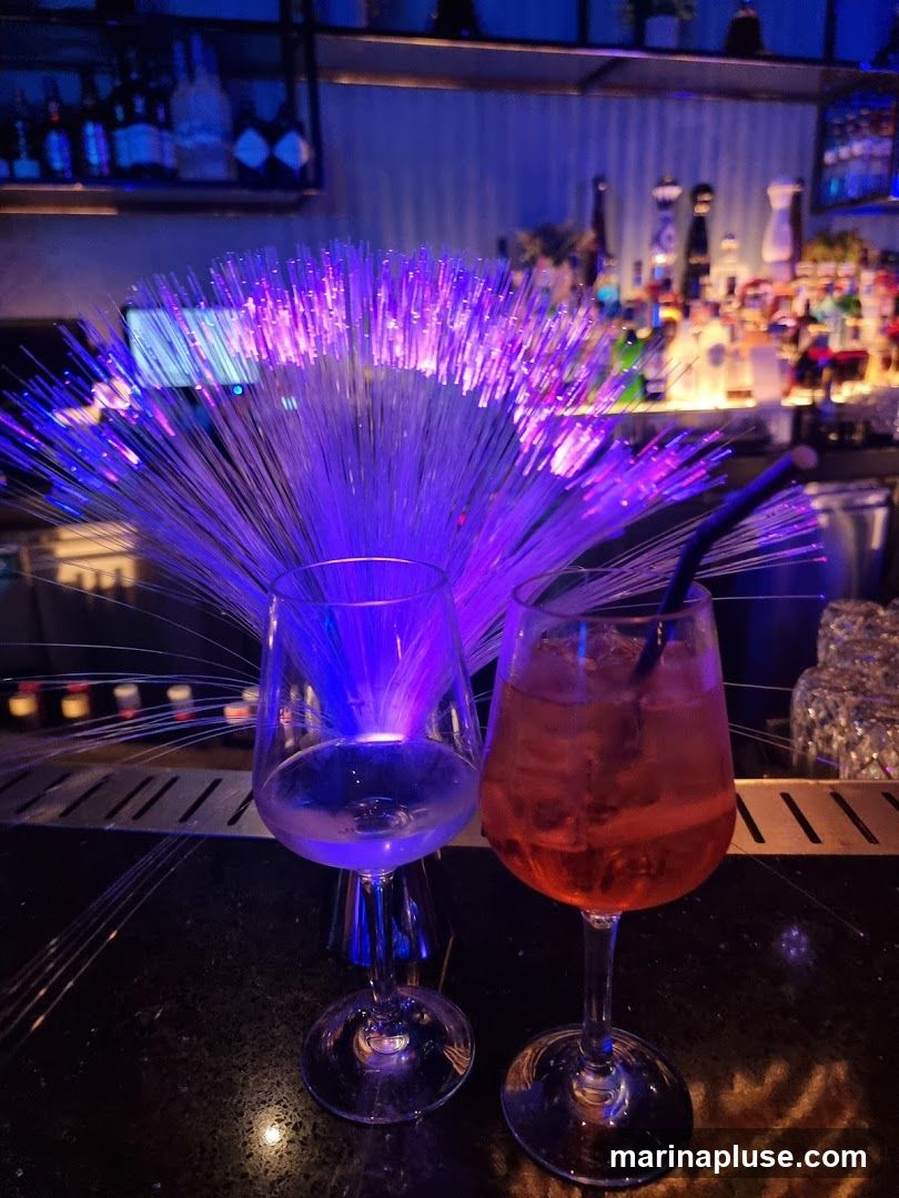 Two wine glasses are on a bar counter. One glass is empty and the other contains a red cocktail with ice and a straw. Behind the glasses, a fiber optic lamp glows with purple and blue light. The background shows a bar with shelves stocked with bottles of alcohol.