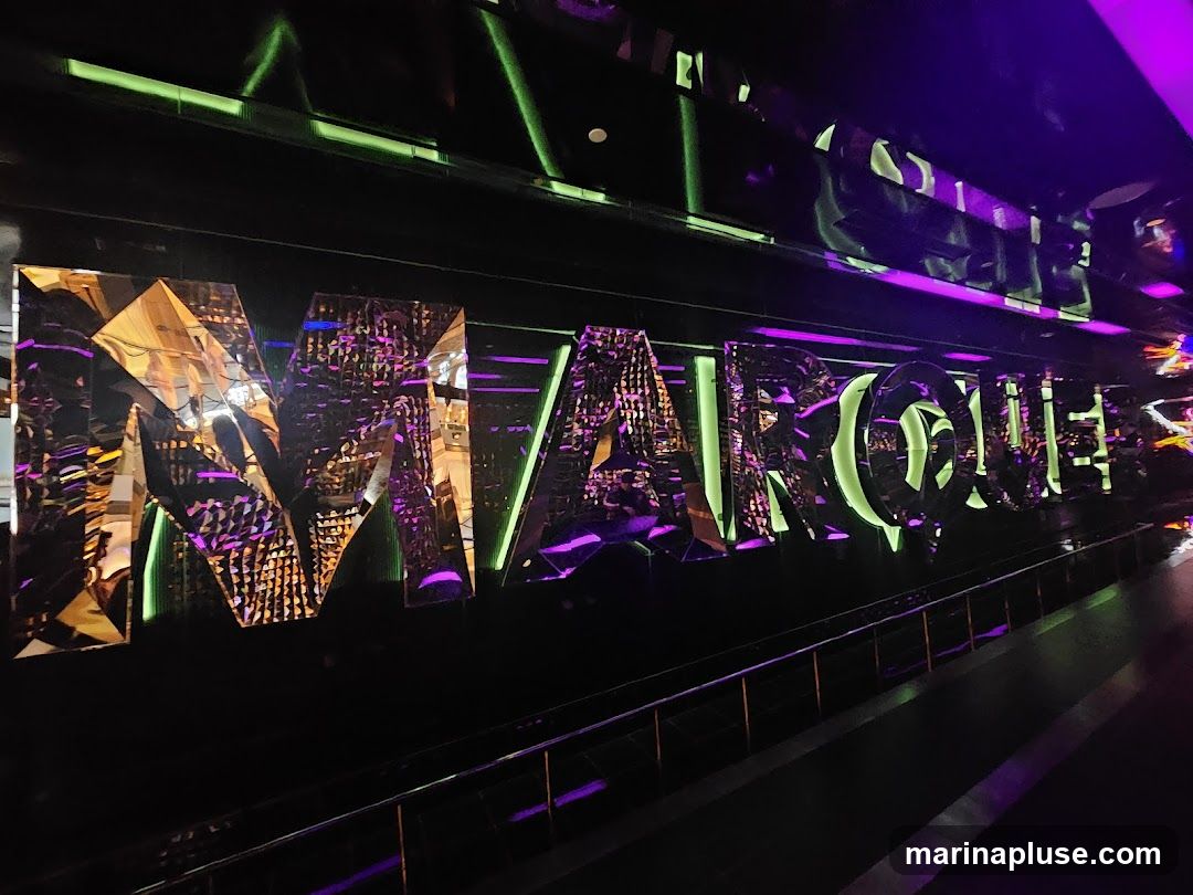 A large, reflective sign spelling out "MARQUEE" is illuminated with green and purple lights. The letters are three-dimensional and appear to be made of a mirrored material, reflecting the surrounding lights and interior. The sign is mounted on a dark background, with a railing visible in the foreground.